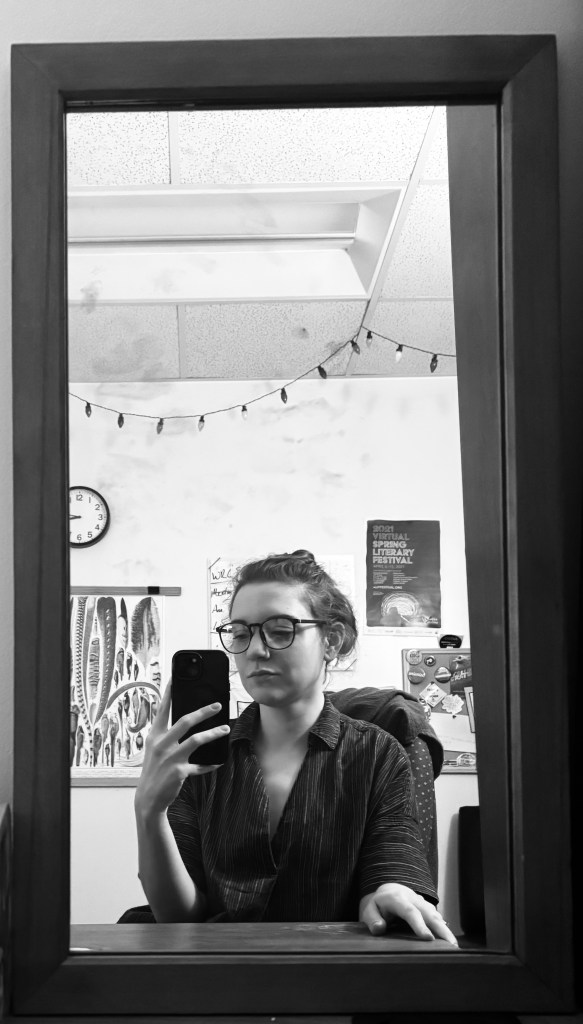 Em takes a black & white cell phone selfie in a rectangular mirror while sitting at their office desk